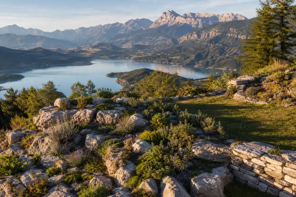 Jardin en terrasse à Chorges sur le versant sud, massifs de rocaille et pelouse basse, lac de Serre-Ponçon scintillant en contrebas, crêtes du Piolit à l'horizon, cadrage panoramique en lumière chaude.