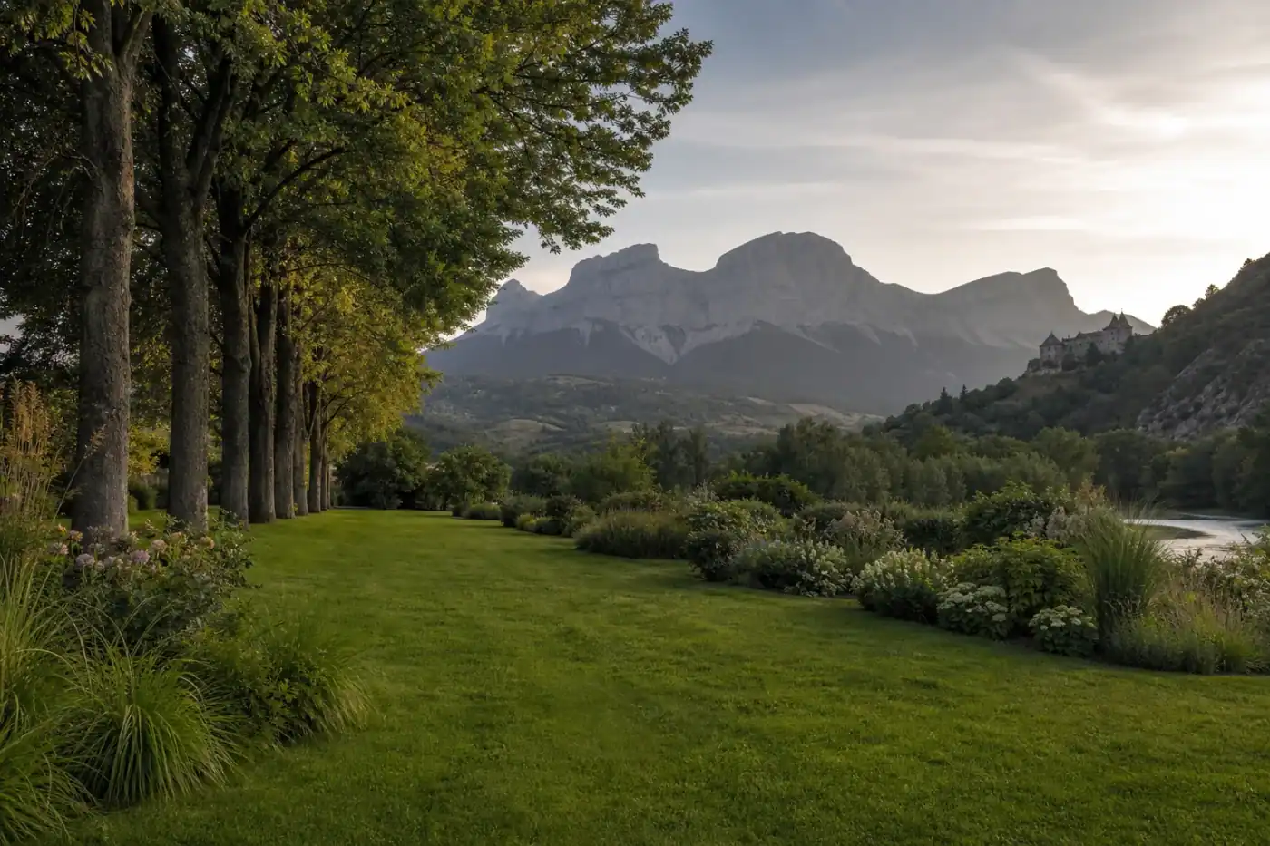 Jardin de plaine à Tallard au bord de la Durance, pelouse dense et massifs fournis, alignement d'arbres, château en silhouette au loin, lumière douce de fin de journée, cadrage horizontal ouvert.