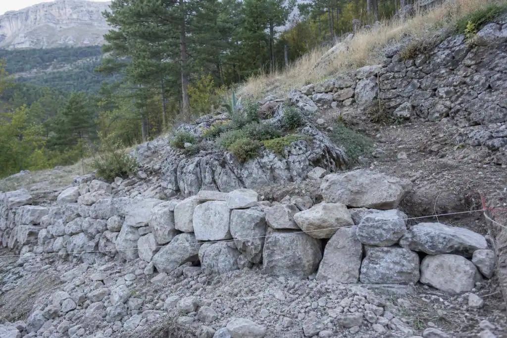 Restanque de pierre sèche en cours de montage sur un terrain en pente, roche claire du Dévoluy, cordeau tendu, massif de rocaille amorcé au-dessus.