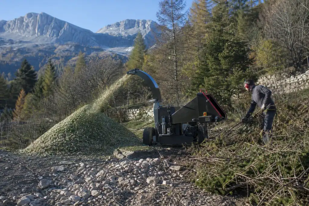 Broyeur de végétaux en action en bord de terrain de montagne, copeaux frais en tas, ronciers coupés en attente, sol caillouteux au premier plan.