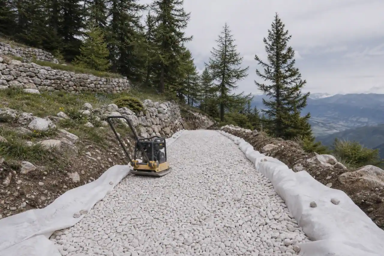 Coupe d'une allée en construction : géotextile et grave concassée en roche claire dans le décaissement, plaque vibrante posée, sol caillouteux gapençais autour.