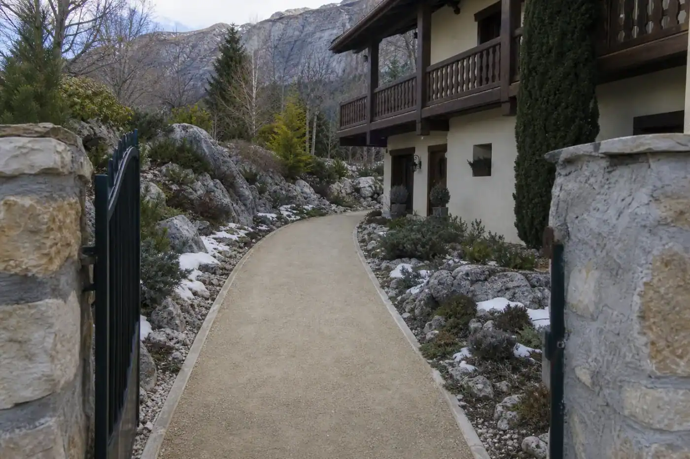 Chemin piéton en stabilisé clair reliant un portail à une entrée de maison au-dessus de Gap, bordures nettes, jardin de rocaille et fonte de neige résiduelle sur les côtés.