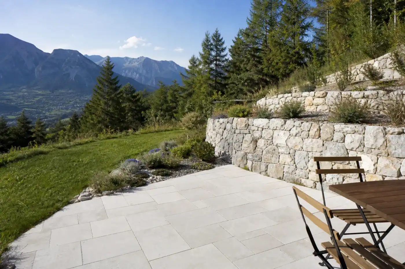 Terrasse en pierre claire fraîchement posée sur un coteau à Gap, calée dans une restanque, ouverte sur une pelouse et les sommets en fond, table et deux chaises simples, fin d'après-midi lumineuse d'altitude.