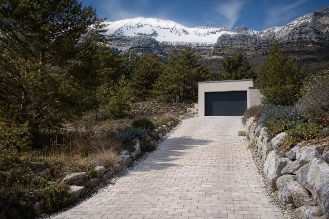 Allée carrossable en pavés clairs menant à un garage sur un terrain en pente à Gap, pente d'écoulement visible sur les côtés, sommets enneigés en fond, lumière d'altitude, aucune personne.