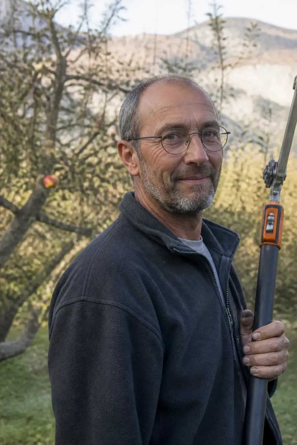 Portrait d'un homme d'une cinquantaine d'années, veste polaire de travail, une perche d'élagage à la main, arbre fruitier de jardin flou derrière lui, sourire discret, cadrage buste, lumière chaude.
