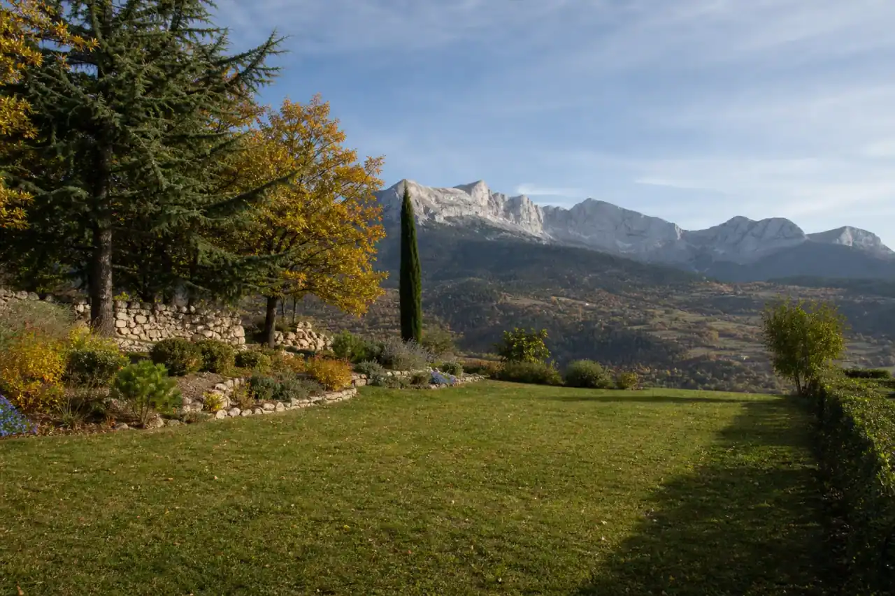 Jardin soigné à Veynes au creux de la vallée du Petit Buëch, pelouse rustique et haie taillée, lumière rasante d'arrière-saison, contreforts du Dévoluy en toile de fond, cadrage large horizontal.